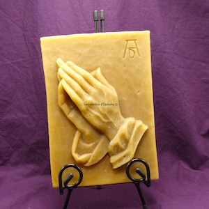 Peut inclure: Une bougie rectangulaire jaune clair avec un relief de mains jointes en prière. La bougie est présentée sur un chevalet en métal noir. Une initiale gravée est visible dans le coin supérieur droit. Le texte "Les abeilles d'Isabelle" est visible.
