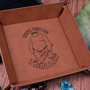 May include: Brown leather dice tray with a black outline of a troll wearing sunglasses and holding a club. The text "They See Me Trollin'" is printed on the tray.