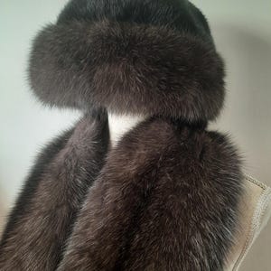 Puede incluir: Un conjunto de gorro y bufanda de piel marrón oscuro. El gorro tiene forma redondeada con un borde esponjoso. La bufanda es larga y también está hecha de piel marrón oscuro. La piel tiene un aspecto suave y texturizado.