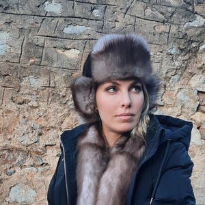 May include: A woman wearing a dark blue jacket with a fur-trimmed hood and a matching fur hat. The hat has ear flaps and is made of a light brown fur.