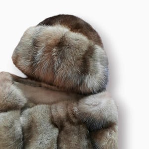 Puede incluir: Un abrigo de piel marrón y gris con un gorro de piel a juego. El abrigo está hecho de piel larga y suave y tiene un diseño clásico.