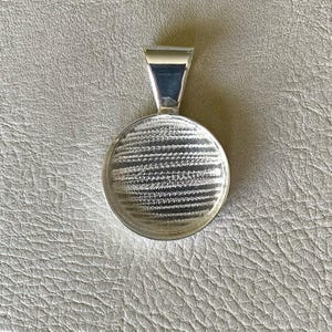May include: A silver-colored pendant with a round, textured face and a rectangular bail. The pendant's surface has a pattern of fine, horizontal lines. The bail is a simple, rectangular shape, and the pendant is set against a neutral background.