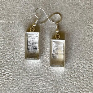 May include: A pair of rectangular earrings with silver-colored hooks. Each earring features a clear, rectangular frame with a white and gray striped design inside. The earrings are displayed against a textured, gray background.