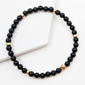 May include: Black beaded bracelet with gold accents. The bracelet is made of small, round black beads with gold spacers and a gold charm.