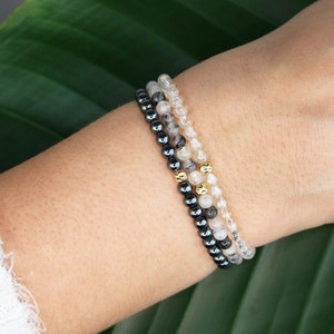 May include: Three bracelets stacked on a wrist. The top bracelet is made of clear quartz beads, the middle bracelet is made of black hematite beads with gold accents, and the bottom bracelet is made of black hematite beads.