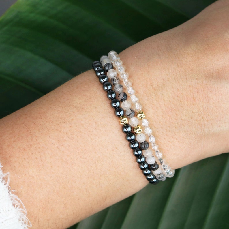 May include: Three bracelets stacked on a wrist. The top bracelet is made of clear quartz beads, the middle bracelet is made of black hematite beads with gold accents, and the bottom bracelet is made of black hematite beads.