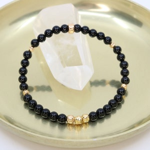 May include: A black beaded bracelet with gold-coloured accents. The bracelet features small, round black beads interspersed with gold-coloured beads and decorative elements. The bracelet is displayed on a gold-coloured tray.