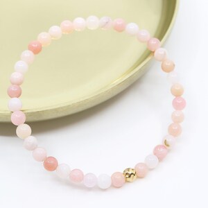 Peut inclure: Un bracelet délicat en pierres précieuses roses et blanches avec un accent de perle dorée. Le bracelet est composé de petites perles rondes et est parfait pour être superposé avec d'autres bracelets.