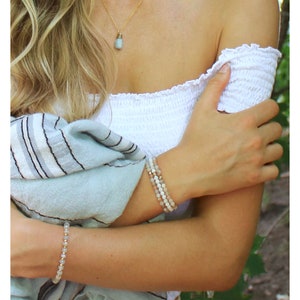 May include: Two beaded bracelets on a woman's wrist. The top bracelet is made of light pink and white beads, while the bottom bracelet is made of white beads with gold accents.
