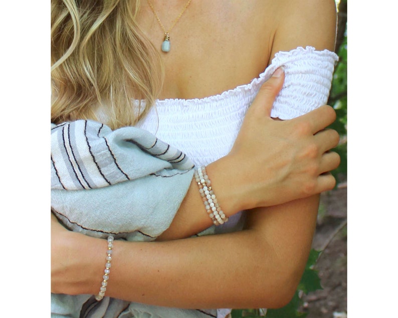 May include: Two beaded bracelets on a woman's wrist. The top bracelet is made of light pink and white beads, while the bottom bracelet is made of white beads with gold accents.