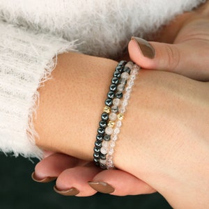 May include: A set of three bracelets featuring black hematite beads, clear quartz beads, and gold beads. The bracelets are stacked on a person's wrist.
