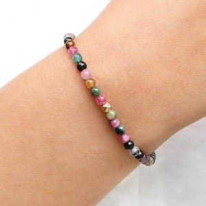 May include: A multi-coloured gemstone bracelet with pink, green, black and gold beads. The bracelet is on a person's wrist.