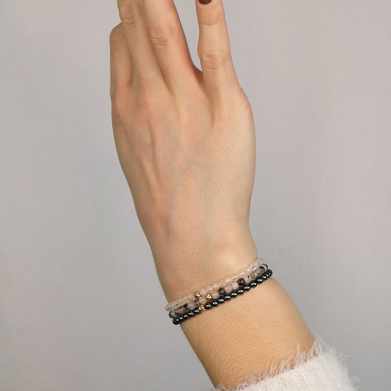May include: Three stacked bracelets on a wrist. The top bracelet is made of clear quartz beads, the middle bracelet is made of rose quartz beads, and the bottom bracelet is made of black hematite beads.