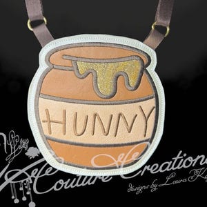 May include: A brown and tan crossbody bag shaped like a honey pot with the word HUNNY embroidered in brown. The bag features a gold glitter applique representing dripping honey, a white stitched border, and an adjustable brown fabric shoulder strap.