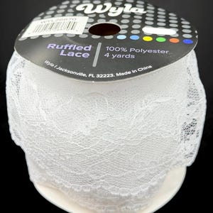 May include: A roll of white ruffled lace made of 100% polyester. The label reads "Ruffled Lace" and "4 yards". The lace is tightly wound on a white cardboard spool. The brand name "Wyla" is printed on the label.