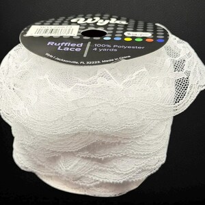 May include: A roll of white ruffled lace. The label reads "Ruffled Lace" and "100% Polyester, 4 yards". The lace is tightly wound and has a delicate, textured appearance. The spool is set against a black background.