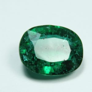 May include: Oval-shaped, faceted emerald gemstone. The gem is a deep, rich green color with internal inclusions, set against a white background. The cut of the stone allows light to reflect, creating a sparkling effect. A beautiful piece for jewelry.