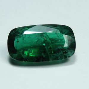 May include: A faceted, emerald-green gemstone with a rectangular cushion cut. The gem has a high degree of clarity and brilliance, reflecting light across its many facets. The stone is set against a white background, highlighting its rich color and shape.