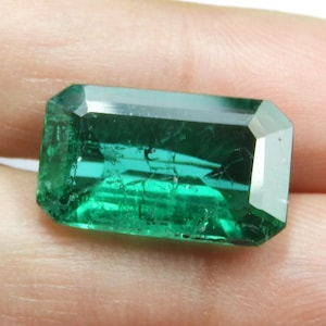 May include: A rectangular, faceted emerald gemstone with a vibrant green hue. The gem's surface reflects light, showcasing its clarity and internal features. The emerald is held in a hand, highlighting its size and shape.