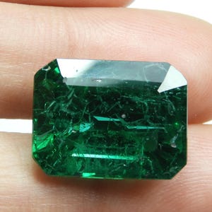 May include: A faceted, emerald-green gemstone with a rectangular shape and cut corners. The gem has internal inclusions and a reflective surface, held between fingers against a white background.
