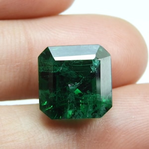 May include: A faceted, emerald-cut gemstone with a deep, rich green color. The gem is held between fingers, showcasing its clarity and internal structure. The cut highlights the stone's brilliance and depth, making it appear luminous.