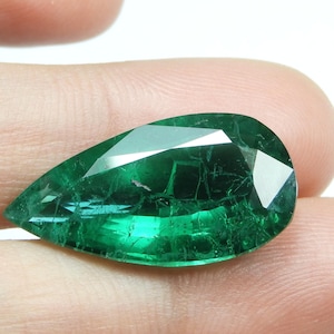 May include: A faceted, pear-shaped emerald gemstone. The gem is a deep, vibrant green color with visible internal inclusions. The cut of the stone creates multiple reflective surfaces.