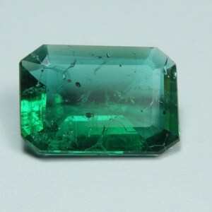 May include: A rectangular, emerald-cut gemstone with a deep green hue. The gem has a transparent appearance with internal inclusions and a polished surface. The facets reflect light, creating a brilliant display. This gemstone is suitable for jewelry.