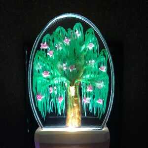 May include: A green willow tree with pink flowers nightlight. The tree is encased in a clear oval-shaped glass. The nightlight has a white base.