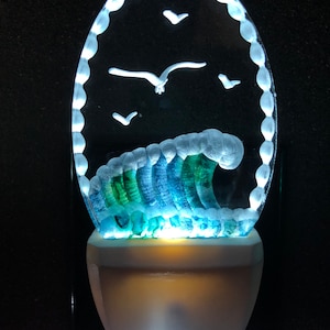 Puede incluir: Una luz de noche decorativa con un diseño de vidrio tallado de una ola y gaviotas. La ola es azul y verde, con detalles en blanco. La luz de noche tiene una base blanca y un borde brillante.