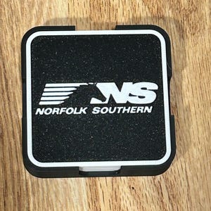 May include: A black square coaster with a white border. The coaster features the white text "NS" above the words "NORFOLK SOUTHERN" in white. The coaster is on a wooden surface.