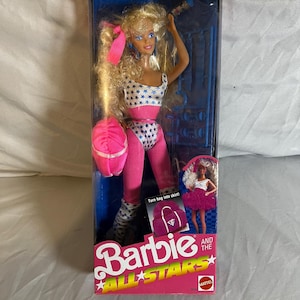 May include: A Barbie All Stars doll in its original packaging. The doll is wearing a white leotard with blue stars, a pink belt, and pink leggings. The packaging includes the text "Aerobics star to party superstar".