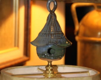 May include: A ceramic birdhouse lamp finial in shades of blue, green, and brown. The finial is attached to a brass lamp harp.