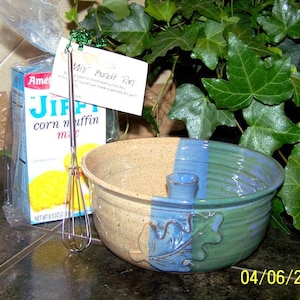 May include: A blue and green ceramic mixing bowl with a built-in whisk holder. The bowl has a leaf design and is sitting on a counter next to a box of Jiffy corn muffin mix and a whisk.