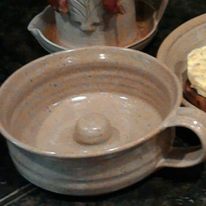 May include: A beige ceramic donut pan with a handle. The pan has a raised center in the middle and is glazed with a light brown glaze.