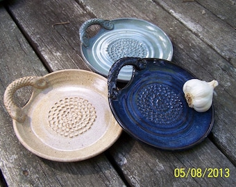 Pode incluir: Três pratos de cerâmica para ralar alho com alças. Um é bege, outro azul e outro azul claro. Um bulbo de alho inteiro está no prato azul.
