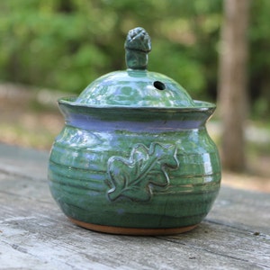 May include: A green ceramic jar with a lid and an oak leaf design. The jar has a light blue glaze and a brown base.