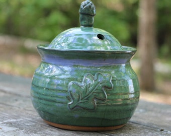 May include: A green ceramic jar with a lid and an oak leaf design. The jar has a light blue glaze and a brown base.