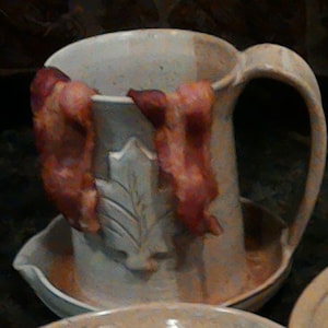 May include: A ceramic pitcher with a leaf design and a handle. Two strips of bacon are hanging from the rim of the pitcher.