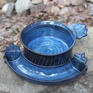 May include: A blue ceramic serving dish with a bowl and a separate dipping dish. The bowl has a leaf-shaped handle on one side. Both pieces have a textured, speckled glaze.