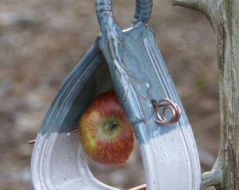 May include: A ceramic bird feeder shaped like a triangle with a white and blue glaze. The feeder has a small opening for birds to access the food and a hook for hanging. A red apple is placed inside the feeder.