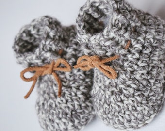 Puede incluir: Un par de botitas de bebé de crochet gris y blanco con lazos de cuero marrón.