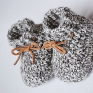 Op de afbeelding: Een paar grijze en witte gehaakte babyschoentjes met bruine leren veters.
