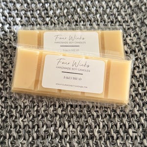 May include: Two rectangular wax melts in clear packaging. Each melt has a label that reads "Four Wicks Handmade Soy Candles BAKED BREAD". The background is a grey knitted fabric. The website address www.fourwicksoycandles.com is also visible.