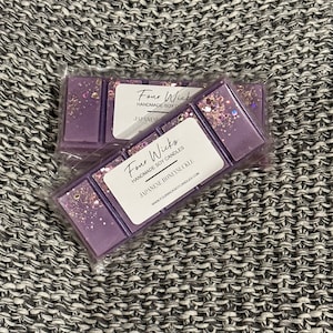 Può includere: Due cubetti di cera di soia viola con glitter. Il cubetto superiore ha un'etichetta che recita "Four Wicks Handmade Soy Candles Japanese Honeysuckle www.fourwickssoycandles.com".