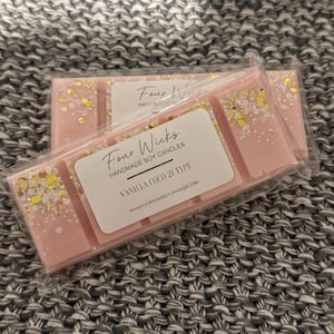 Puede incluir: Un derretimiento de cera rosa y dorado con el texto "Four Wicks Handmade Soy Candles" y "Vanilla Coco 21 Type" en el empaque. El derretimiento de cera está en una envoltura de plástico transparente.