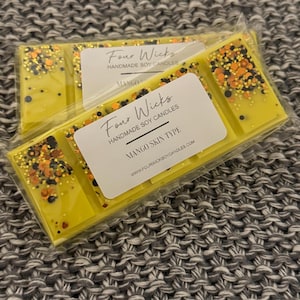 May include: A yellow soy wax melt with black and orange glitter. The melt is labelled "Four Wicks Handmade Soy Candles" and "Mango Skin Type".