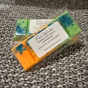 May include: Two wax melts, one green and one orange, with glitter. The wax melts are packaged in clear plastic and labelled "Four Wicks Handmade Soy Candles Basil Lime and Mandarinine" with the website address "www.fourwicksoycandles.com".