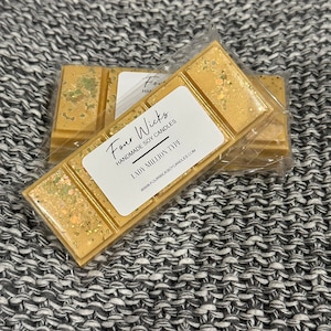 May include: A gold and white soy wax melt with the text "Four Wicks Handmade Soy Candles Lady Million Type" and the website address "www.fourwickssoycandles.com".