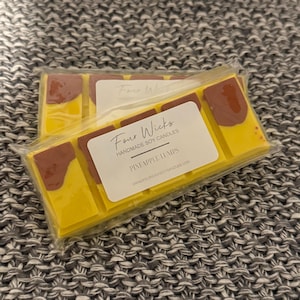 May include: A close-up of a wax melt bar with a yellow and brown design. The bar is labelled "Four Wicks Handmade Soy Candles Pineapple Lumps" and includes the website address "www.fourwickssoyscandles.com".