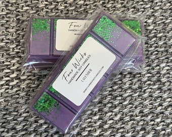 Op de afbeelding: Twee paarse sojawas smelttabletten met groene glitter. De smelttabletten zijn verpakt in doorzichtig plastic en gelabeld met "Four Wicks Handmade Soy Candles Lavender www.fourwicksoycandles.com"